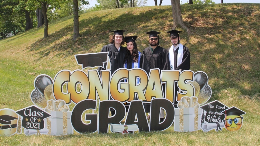 Four graduates in caps and gowns standing among Congrats Grad lawn signs and ornaments