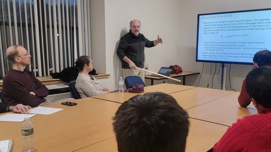 Man holding pointer lectures before and LED screen to a group sitting around a table