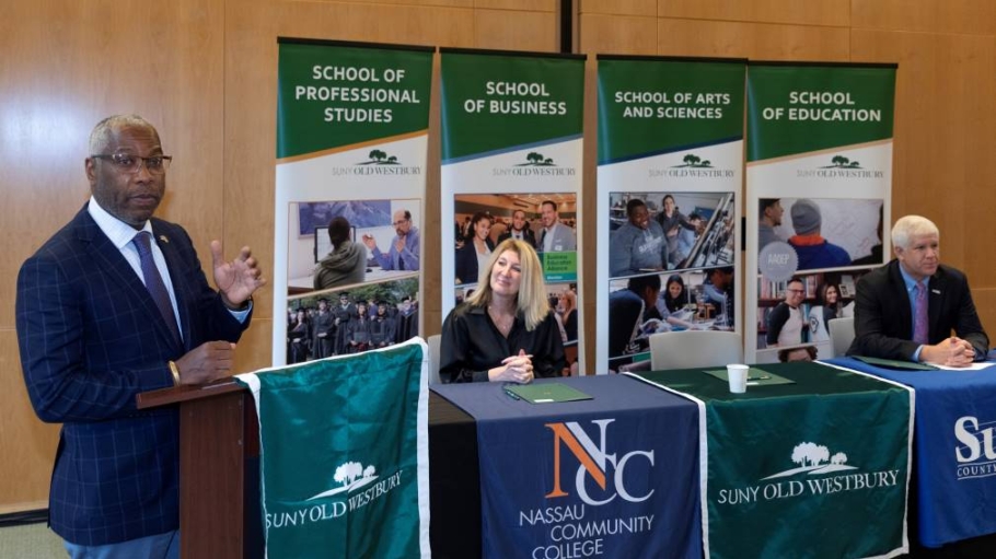 One man at a podium while a man and women are seated a table covered with banners from colleges