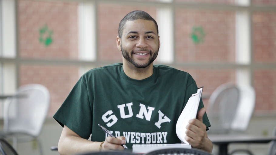 A male student wearing green SUNY Old Westbury shirt sitting at a desk with an open book and smiling at the camera