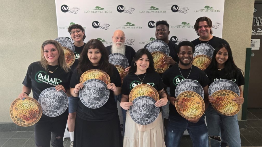 A diverse group of 10 people wearing black tee shirts hold copies of a silver and gold circular award before a step and repeat featuring the college logo and radio station logo