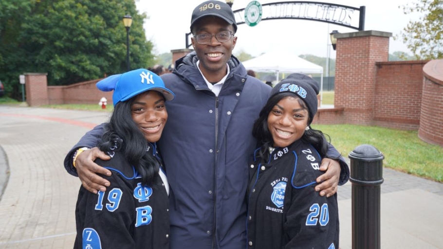 An African American man with a baseball hat and glasses poses with two African American women wearing sorority-oriented jackets