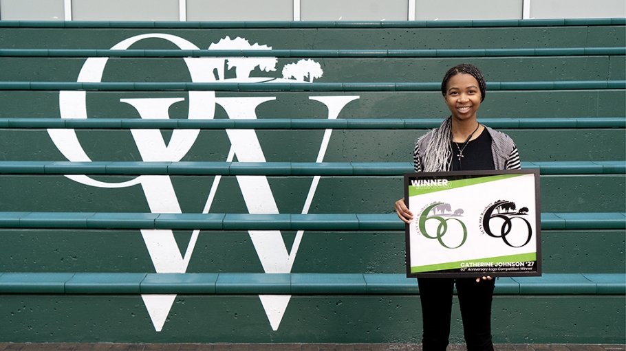 African American young woman stands before a flight of green steps with letters O and W in white while holding a framed print of a logo featuring the number 60 