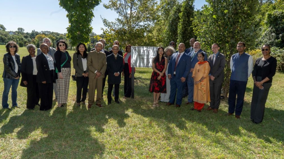 A group of 25 people from diverse backgrounds pose before a stand of trees and a sign reading Ghandi Peace Garden