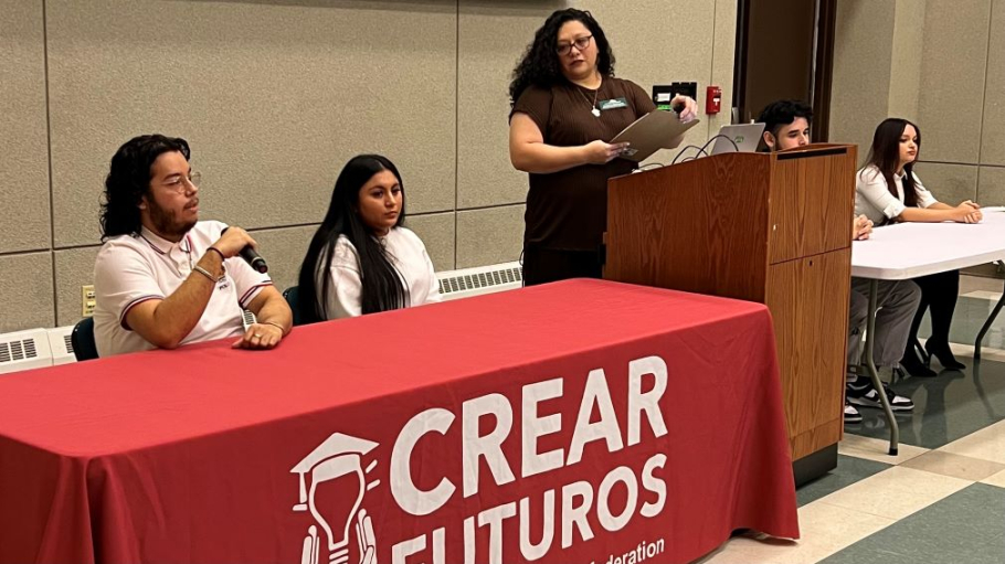 Four students seated at tables with microphones before a diverse group of students