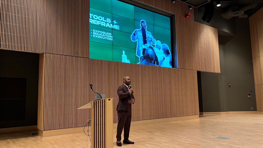 African American man in suit speaks from a stage with a large presentation screen behind him