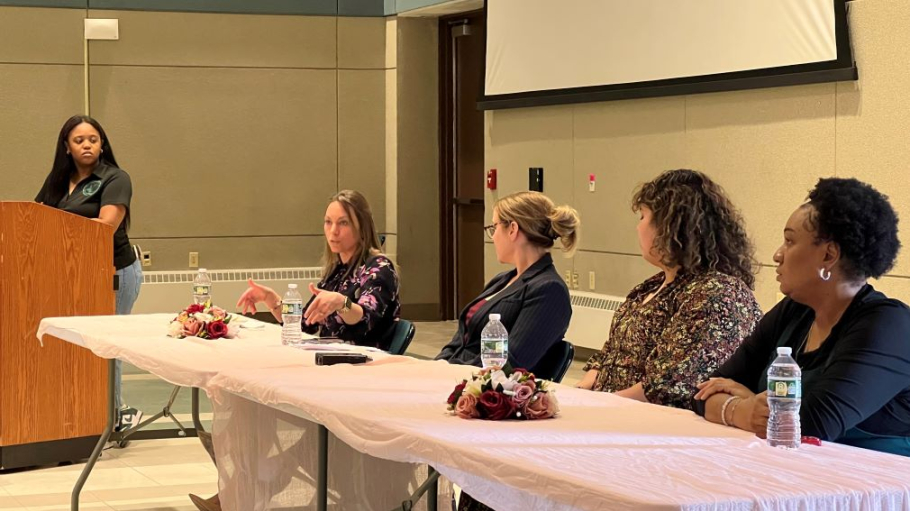 An African American young woman at podium with four other professional women seated at a table