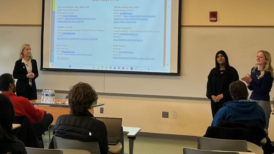 Three women in professional attire speak before a classroom of students with a presentation showing on the room's lightboard