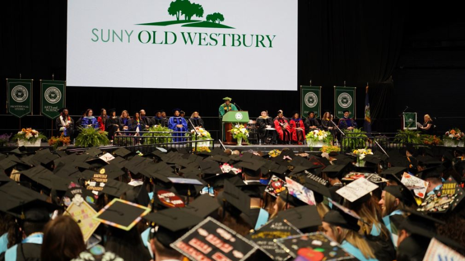 A crowd of graduates wearing decorated mortarboards face a stage set for a commencement ceremony.