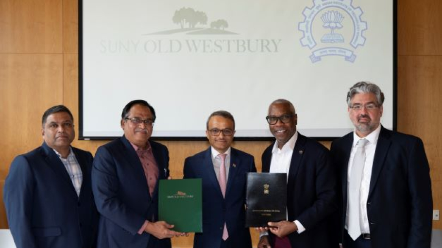 A group of five men from diverse backgrounds pose with two holding folders before a screen showing the logos of the Indian Institute of Technology Bombay and SUNY Old Westbury