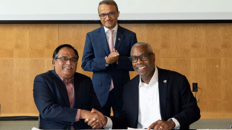 A man of South Asian descent and an African American man shake hands while smiling as another man of South Asian descent looks on