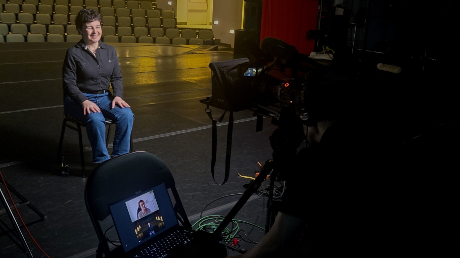 Professor sitting and smiling on chair on Maguire Theater stage while looking towards a camera. Video conference streaming on laptop on chair.