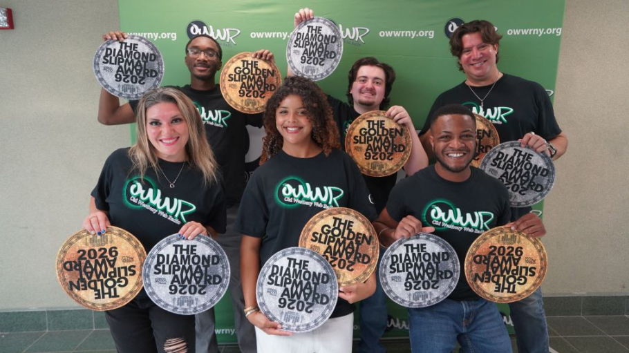 A diverse group of six people wearing black tee shirts standing in front of a step and repeat featuring the radio station logo