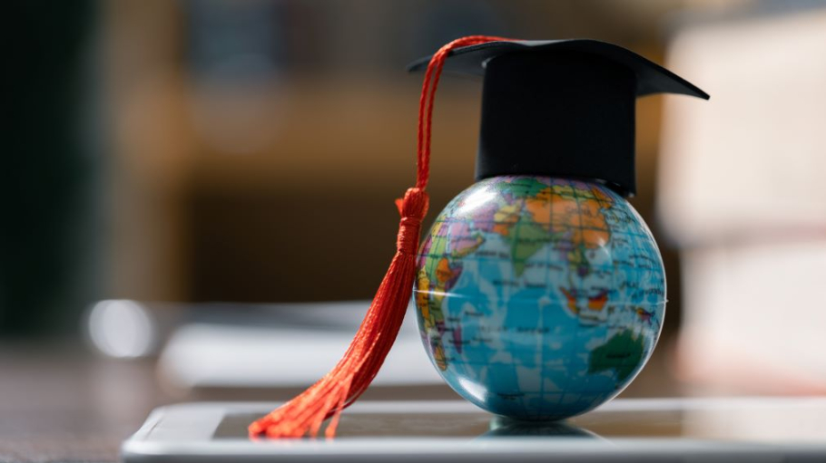Photo illustration featuring a small globe resting on a laptop keyboard while wearing a mortarboard and tassel 