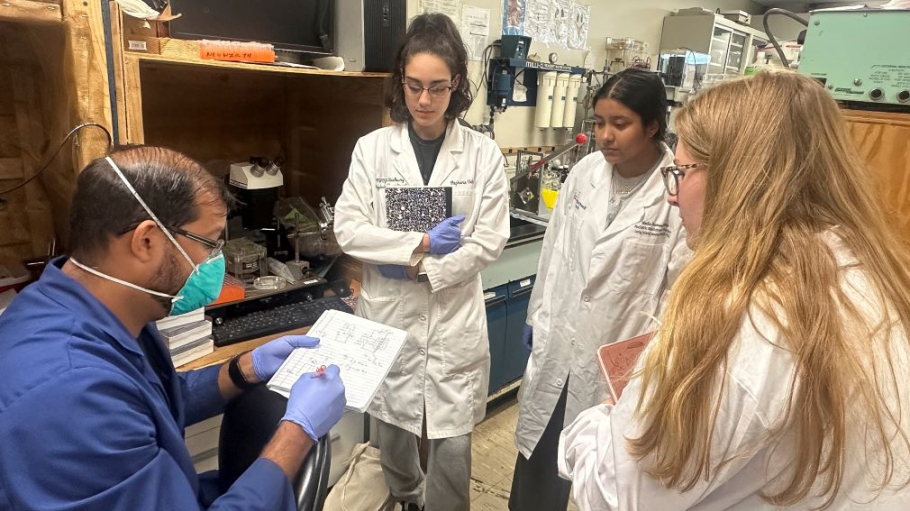 Three young ladies in lab coats work with a professor in a science research lab