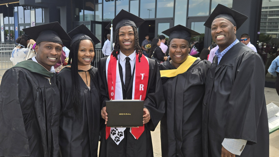 Group of five wearing Commencement regalia standing and smiling together as young man in the middle holds a diploma cover.
