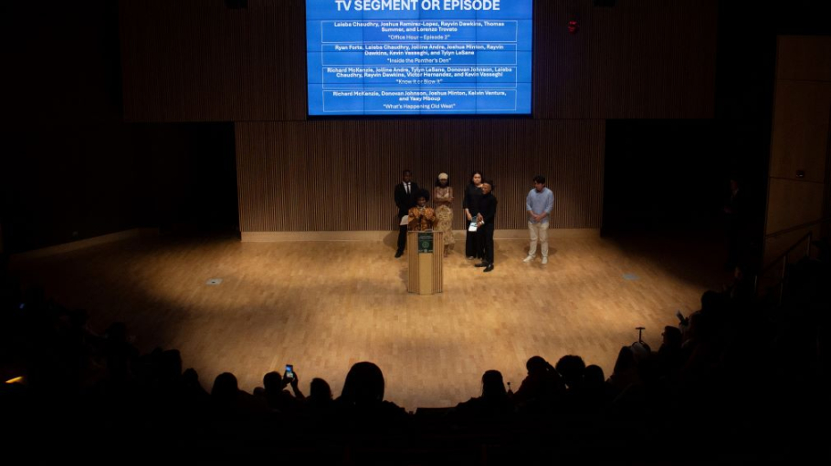Five students on a brightly lit stage before a large audience cast in shadow