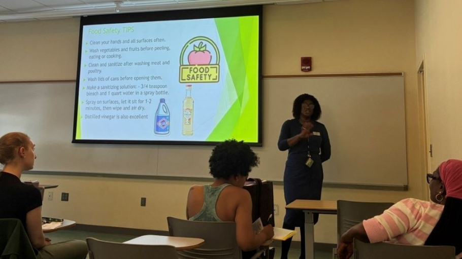 African American woman in professional dress lectures to a group of women while standing in front of a screen that shows a slide on food safety