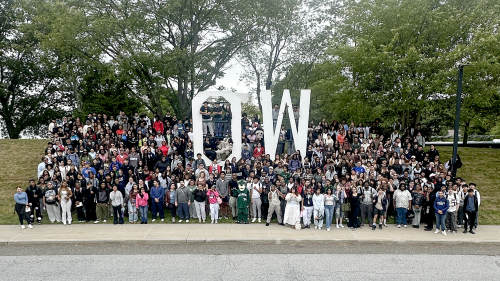 A crowd of hundreds of students gathered together for a group photo in front of large white OW letters on the SUNY Old Westbury campus