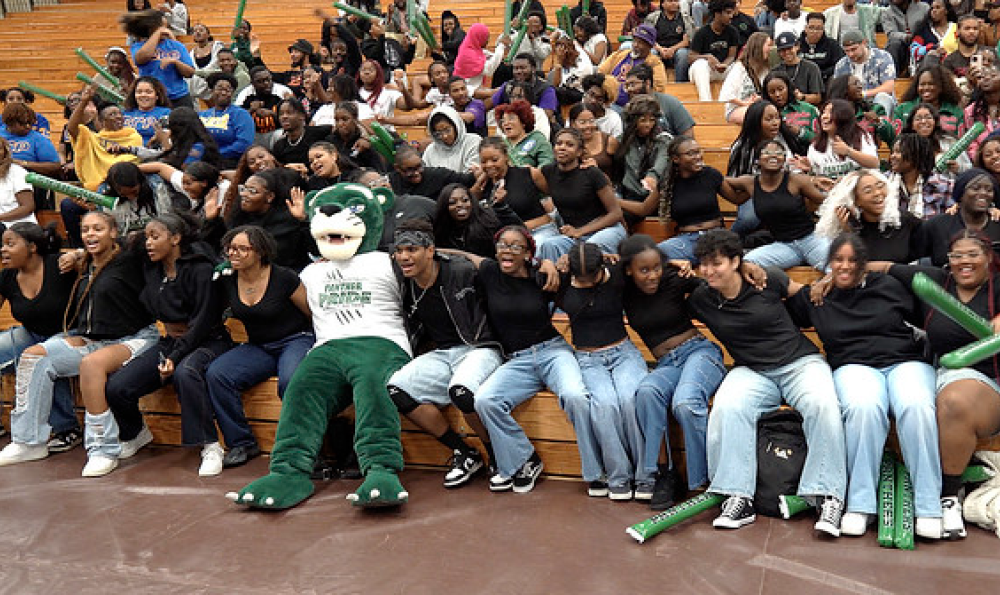 Large group of students sitting in bleachers with a green panther mascot