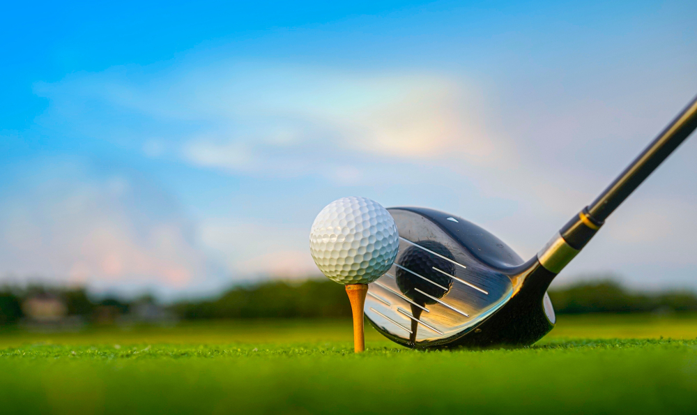 A golf ball on a tee next to a driver club on a sunny green field
